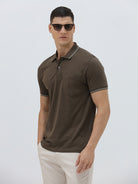 A man wearing a dark brown short-sleeve polo shirt with contrasting light gray trim on the collar and sleeve cuffs, paired with off-white trousers, against a plain light gray background.