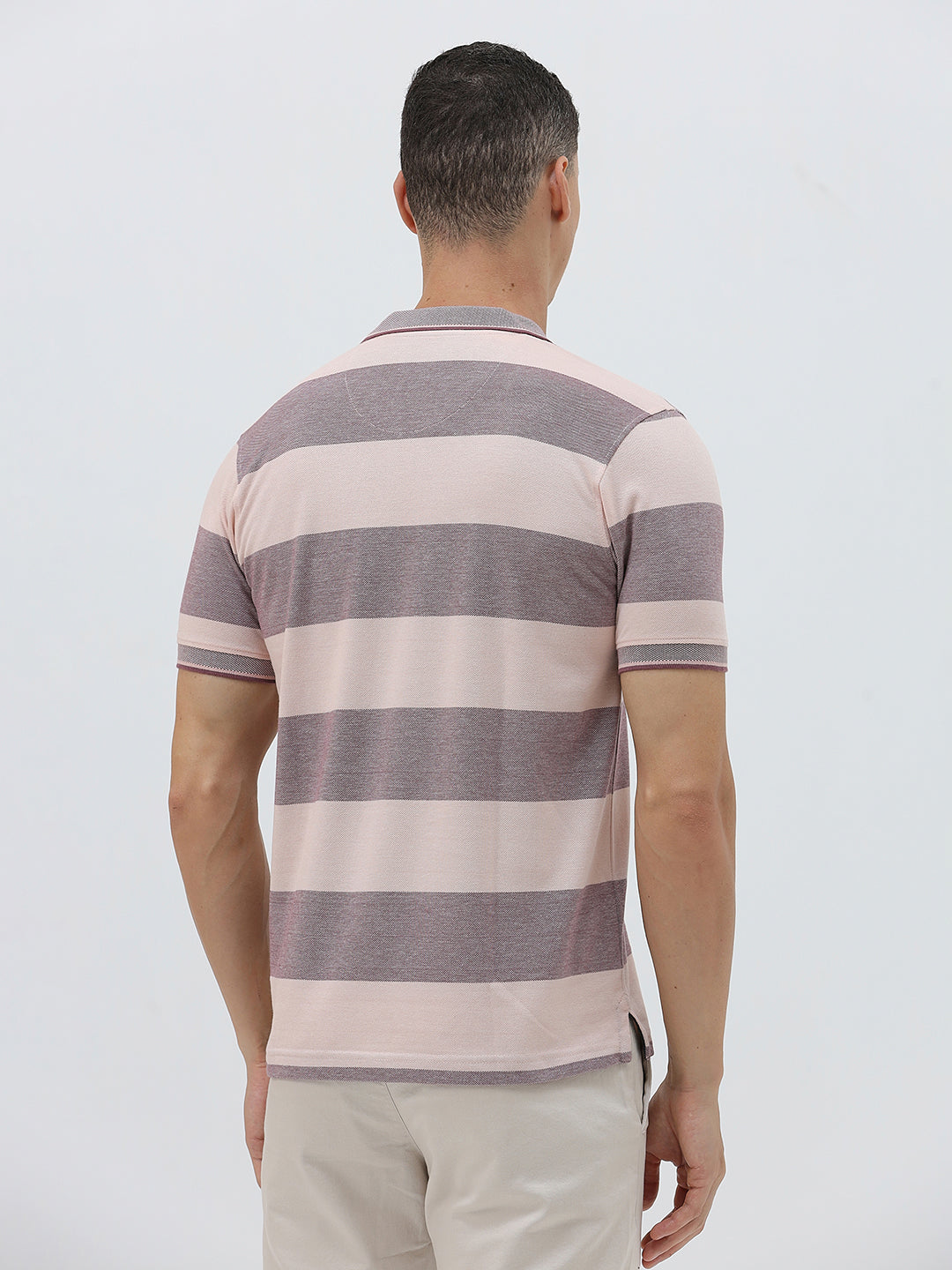 A man wearing a short-sleeve polo shirt with horizontal stripes in dusty pink and gray, paired with light beige trousers, against a plain light gray background.