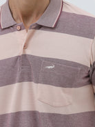 A man wearing a short-sleeve polo shirt with horizontal stripes in dusty pink and gray, paired with light beige trousers, against a plain light gray background.