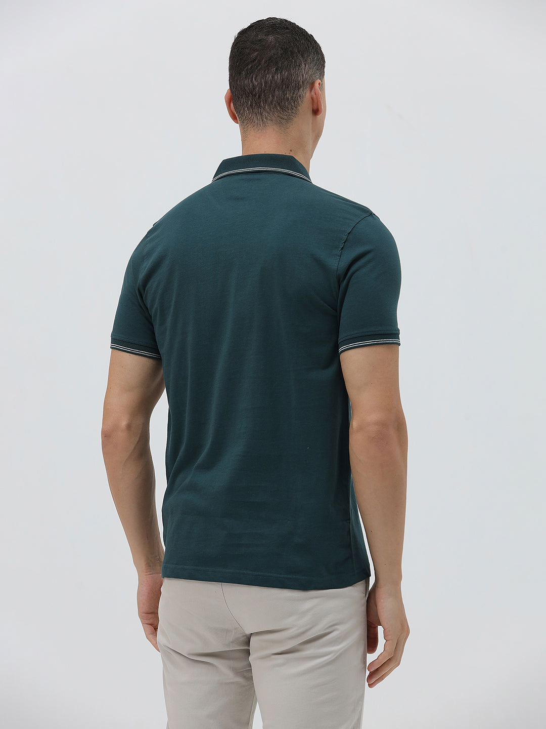 A man wearing a dark teal-green short-sleeve polo shirt with contrasting light gray piping on the collar and sleeve cuffs, paired with light beige trousers, against a plain light gray studio background.