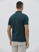 A man wearing a dark teal-green short-sleeve polo shirt with contrasting light gray piping on the collar and sleeve cuffs, paired with light beige trousers, against a plain light gray studio background.
