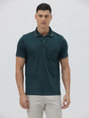 A man wearing a dark teal-green short-sleeve polo shirt with contrasting light gray piping on the collar and sleeve cuffs, paired with light beige trousers, against a plain light gray studio background.