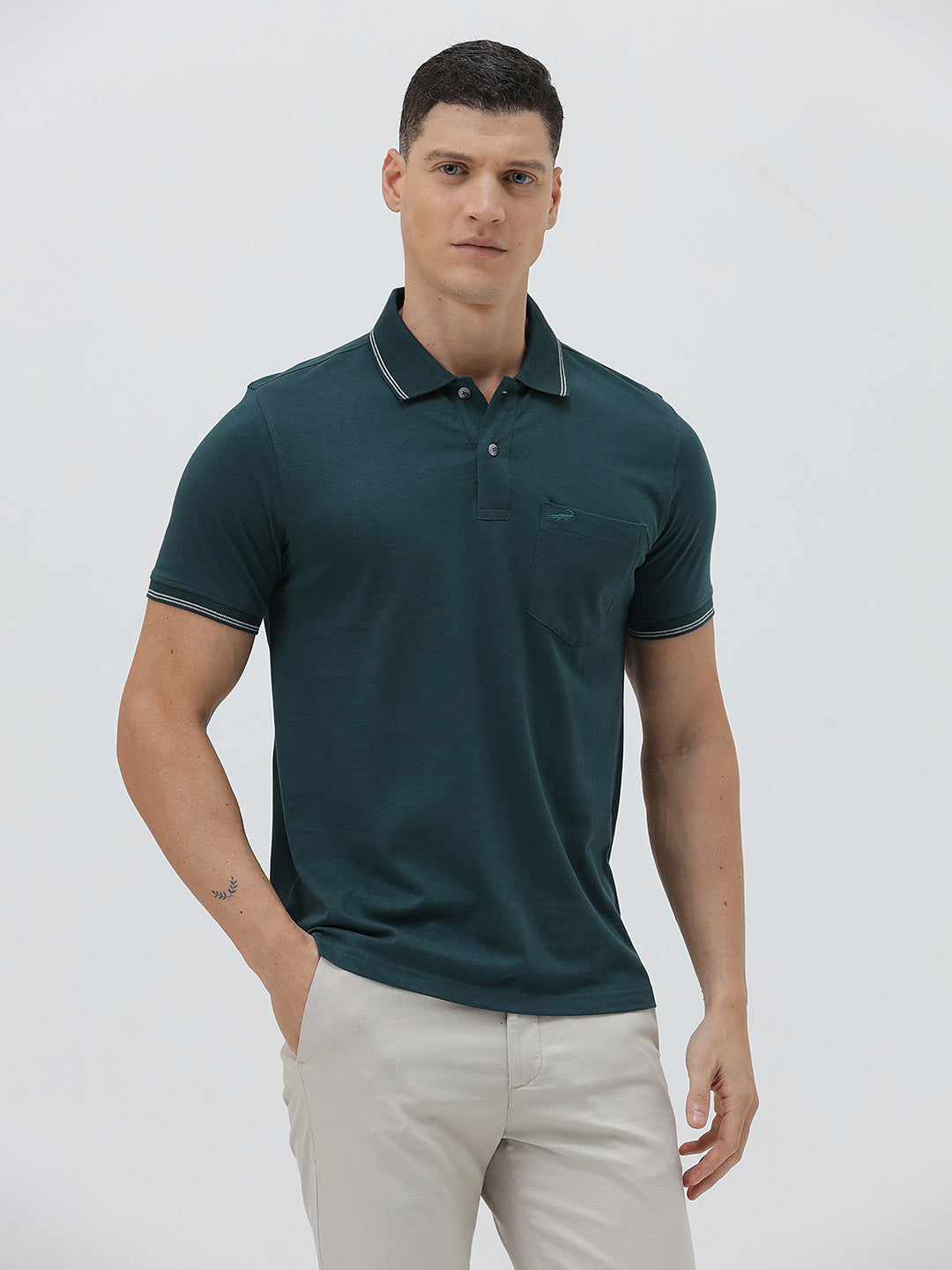 A man wearing a dark teal-green short-sleeve polo shirt with contrasting light gray piping on the collar and sleeve cuffs, paired with light beige trousers, against a plain light gray studio background.