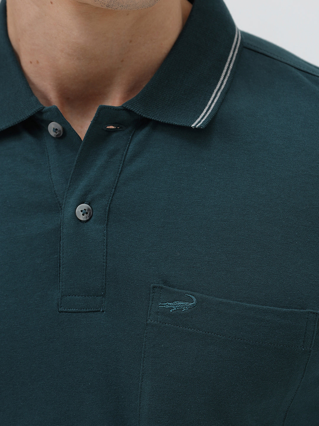 A man wearing a dark teal-green short-sleeve polo shirt with contrasting light gray piping on the collar and sleeve cuffs, paired with light beige trousers, against a plain light gray studio background.