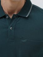 A man wearing a dark teal-green short-sleeve polo shirt with contrasting light gray piping on the collar and sleeve cuffs, paired with light beige trousers, against a plain light gray studio background.