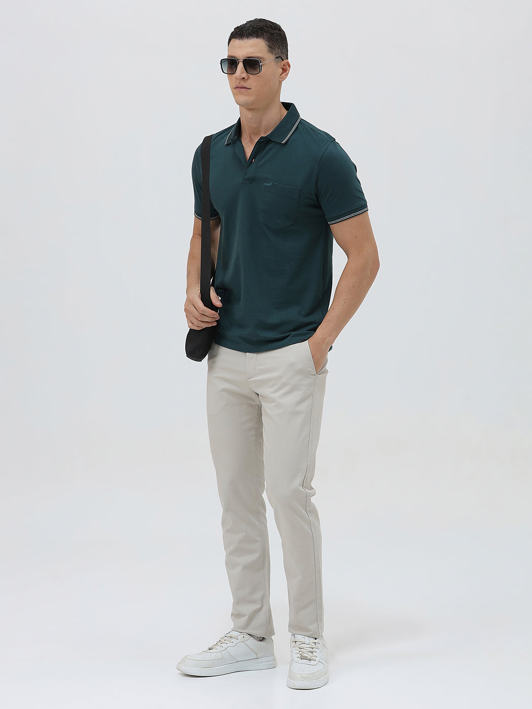 A man wearing a dark teal-green short-sleeve polo shirt with contrasting light gray piping on the collar and sleeve cuffs, paired with light beige trousers, against a plain light gray studio background.