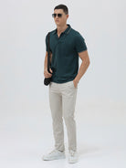 A man wearing a dark teal-green short-sleeve polo shirt with contrasting light gray piping on the collar and sleeve cuffs, paired with light beige trousers, against a plain light gray studio background.
