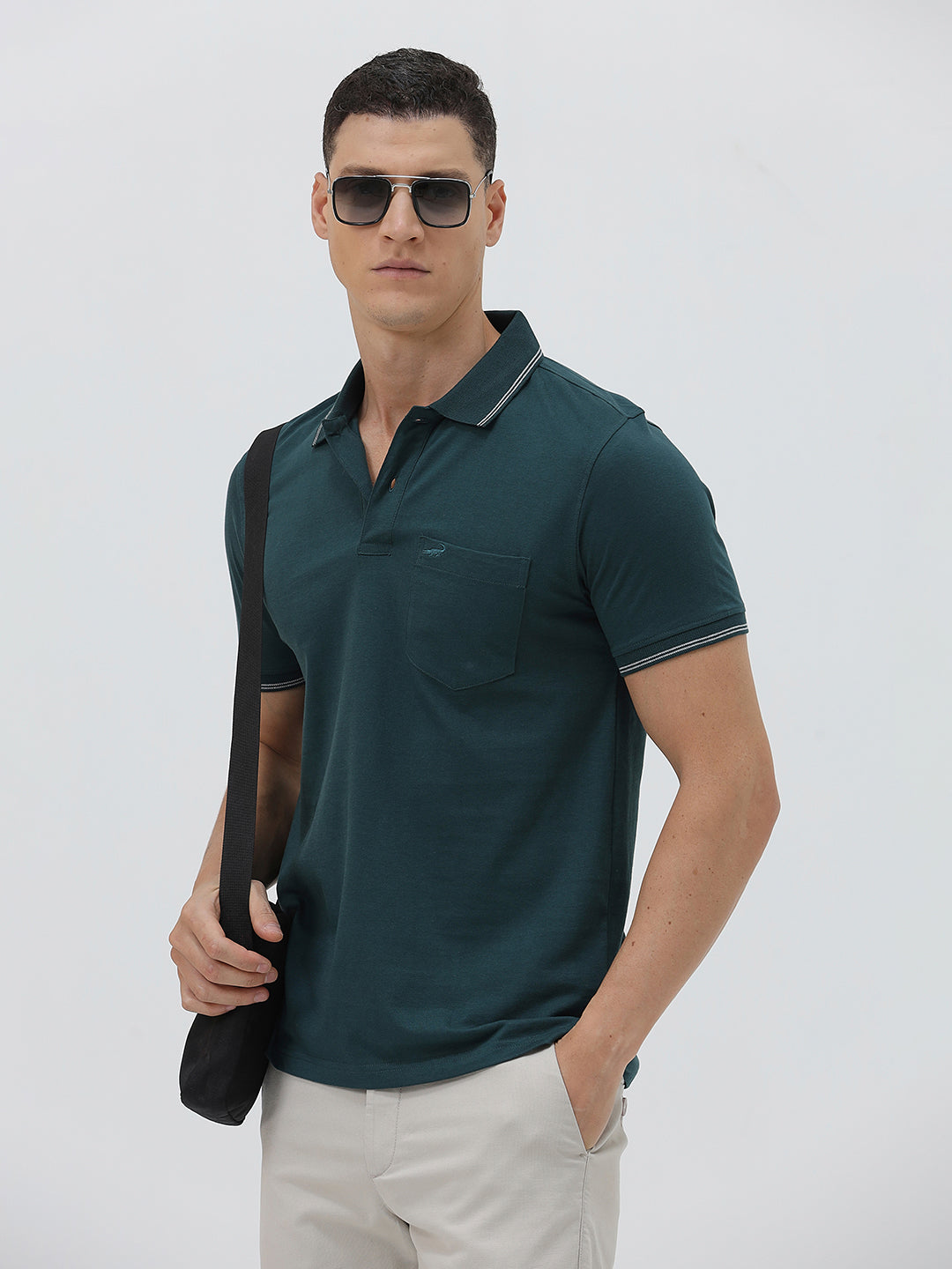 A man wearing a dark teal-green short-sleeve polo shirt with contrasting light gray piping on the collar and sleeve cuffs, paired with light beige trousers, against a plain light gray studio background.