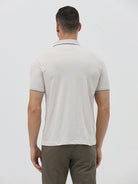 A man wearing a light beige short-sleeve polo shirt with contrasting dark gray piping on the collar and sleeve cuffs, paired with olive green trousers, against a plain light gray background.