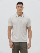 A man wearing a light beige short-sleeve polo shirt with contrasting dark gray piping on the collar and sleeve cuffs, paired with olive green trousers, against a plain light gray background.