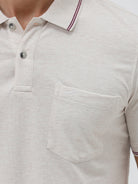 A man wearing a light beige short-sleeve polo shirt with contrasting dark gray piping on the collar and sleeve cuffs, paired with olive green trousers, against a plain light gray background.