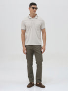A man wearing a light beige short-sleeve polo shirt with contrasting dark gray piping on the collar and sleeve cuffs, paired with olive green trousers, against a plain light gray background.