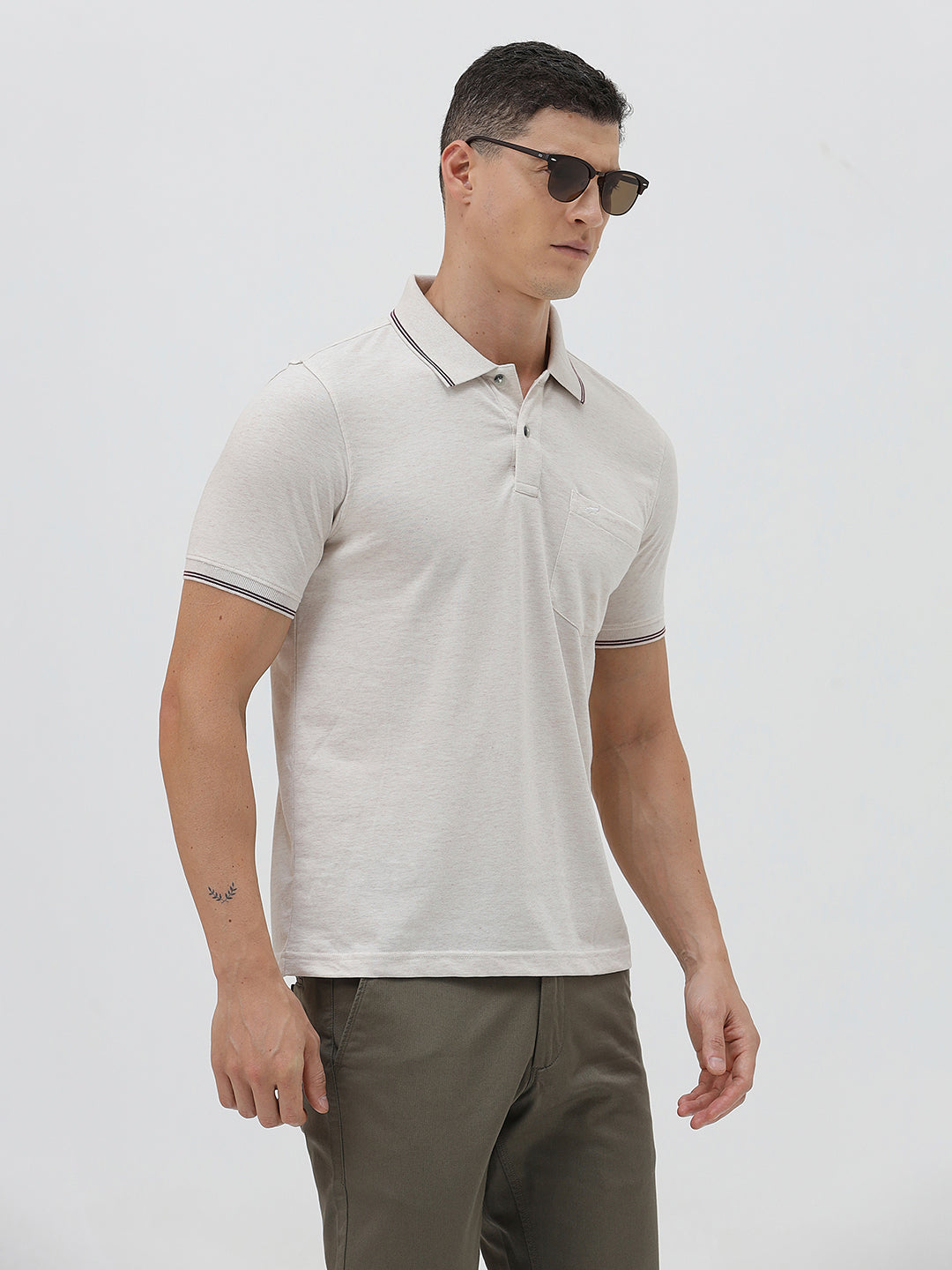 A man wearing a light beige short-sleeve polo shirt with contrasting dark gray piping on the collar and sleeve cuffs, paired with olive green trousers, against a plain light gray background.