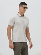 A man wearing a light beige short-sleeve polo shirt with contrasting dark gray piping on the collar and sleeve cuffs, paired with olive green trousers, against a plain light gray background.