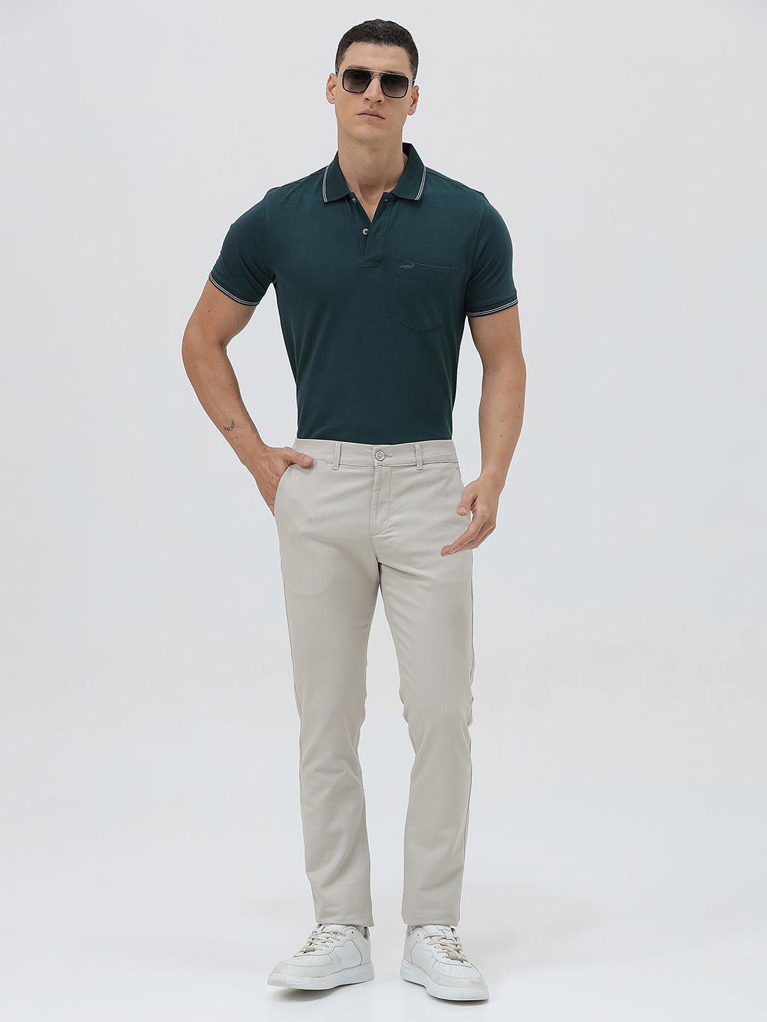 A person wearing slim-fit light beige chinos with a dark green t-shirt and white sneakers, standing against a plain white studio background.