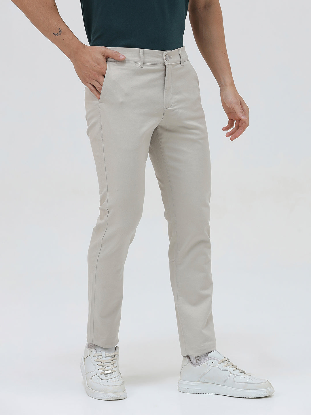 A person wearing slim-fit light beige chinos with a dark green t-shirt and white sneakers, standing against a plain white studio background.