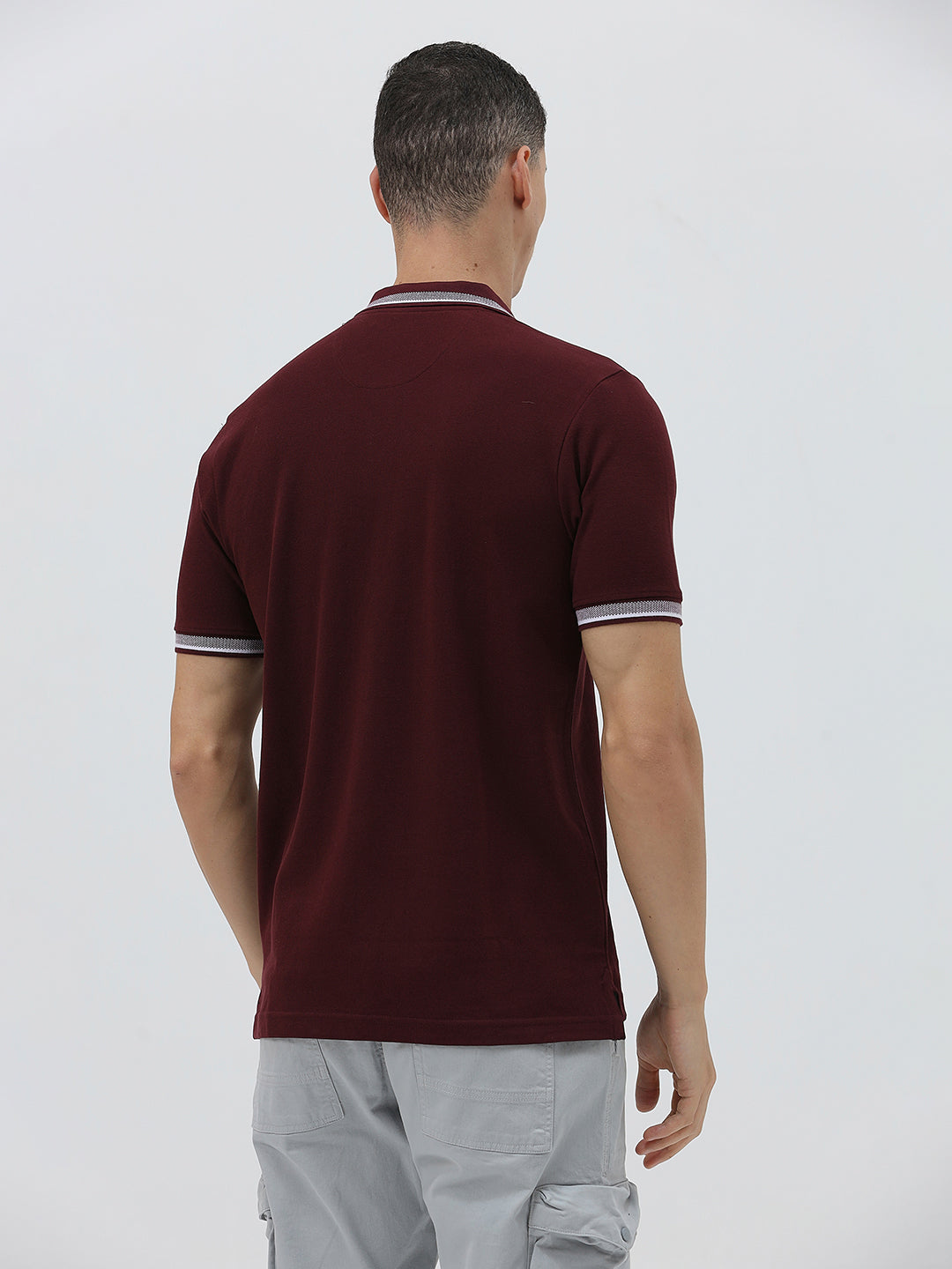 A man wearing a short-sleeve polo shirt in maroon with a contrasting light gray and white striped collar and sleeve cuffs, paired with light gray cargo pants, against a plain light gray background.