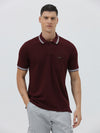 A man wearing a short-sleeve polo shirt in maroon with a contrasting light gray and white striped collar and sleeve cuffs, paired with light gray cargo pants, against a plain light gray background.