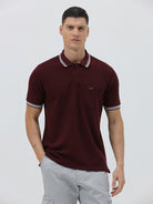 A man wearing a short-sleeve polo shirt in maroon with a contrasting light gray and white striped collar and sleeve cuffs, paired with light gray cargo pants, against a plain light gray background.