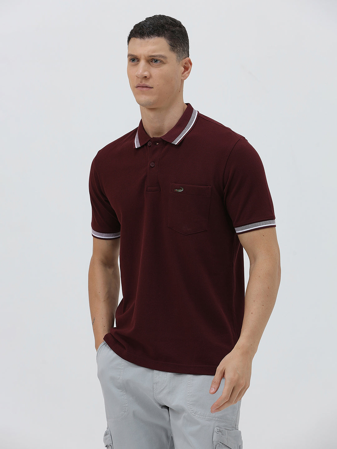 A man wearing a short-sleeve polo shirt in maroon with a contrasting light gray and white striped collar and sleeve cuffs, paired with light gray cargo pants, against a plain light gray background.
