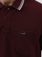 A man wearing a short-sleeve polo shirt in maroon with a contrasting light gray and white striped collar and sleeve cuffs, paired with light gray cargo pants, against a plain light gray background.