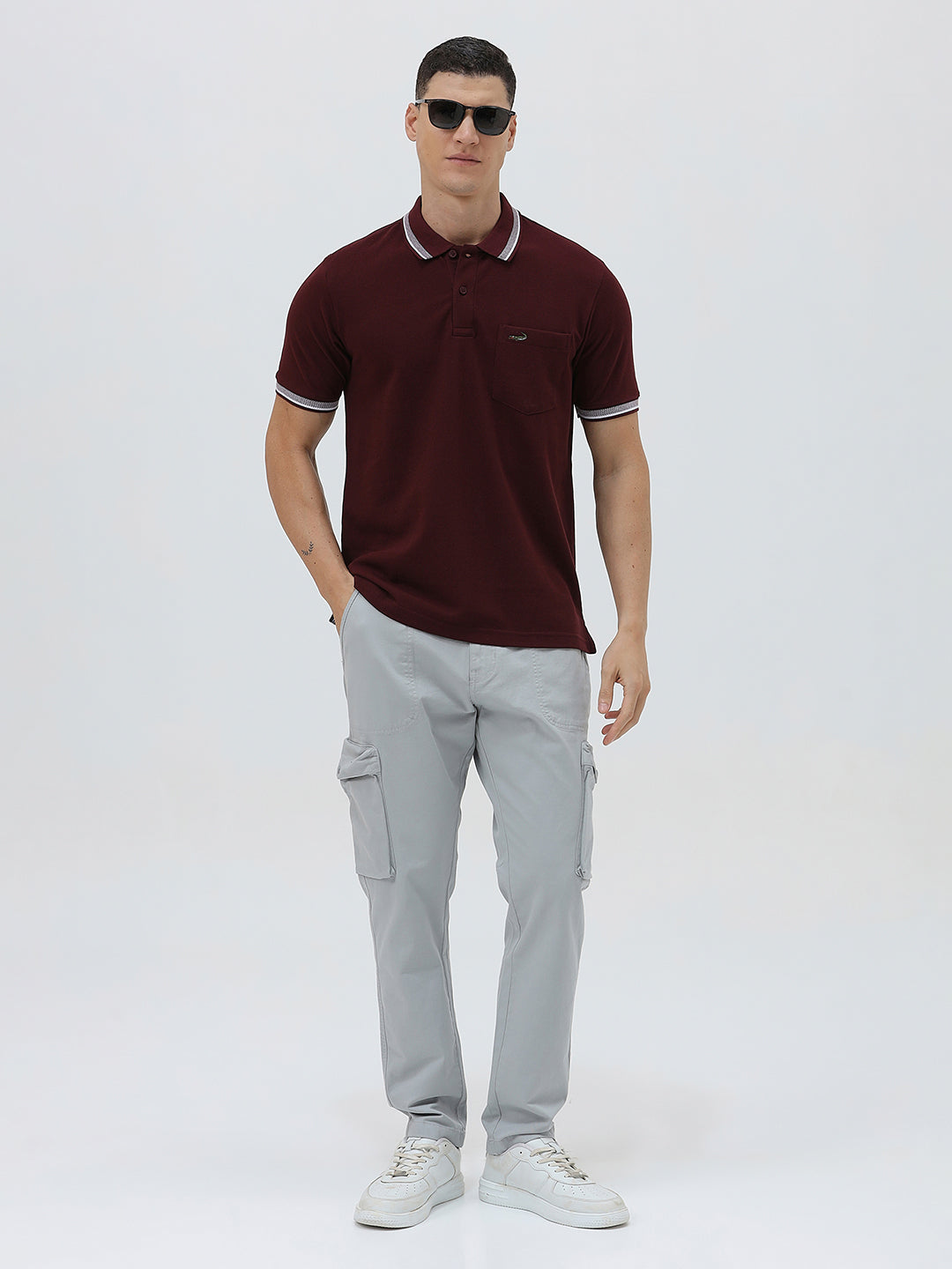 A man wearing a short-sleeve polo shirt in maroon with a contrasting light gray and white striped collar and sleeve cuffs, paired with light gray cargo pants, against a plain light gray background.