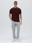 A man wearing a short-sleeve polo shirt in maroon with a contrasting light gray and white striped collar and sleeve cuffs, paired with light gray cargo pants, against a plain light gray background.