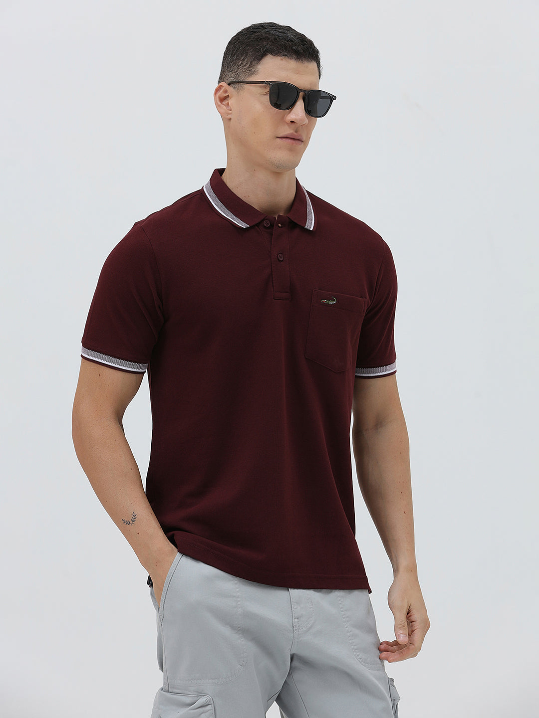 A man wearing a short-sleeve polo shirt in maroon with a contrasting light gray and white striped collar and sleeve cuffs, paired with light gray cargo pants, against a plain light gray background.