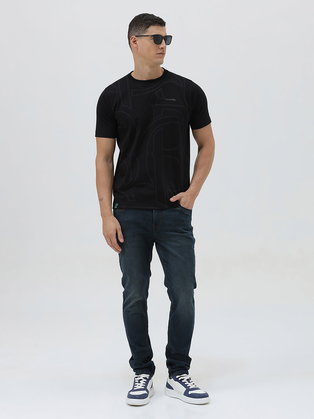 A person wearing dark blue slim-fit denim jeans, paired with a black t-shirt and navy and white sneakers, standing against a plain white studio background.