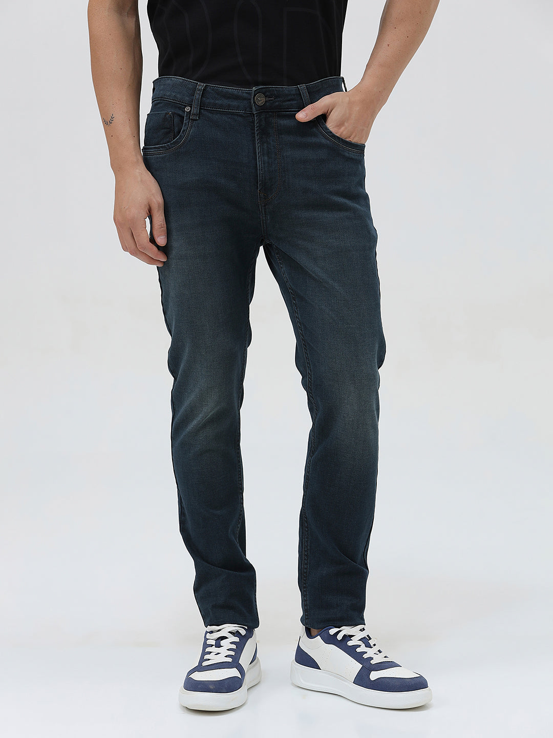 A person wearing dark blue slim-fit denim jeans, paired with a black t-shirt and navy and white sneakers, standing against a plain white studio background.