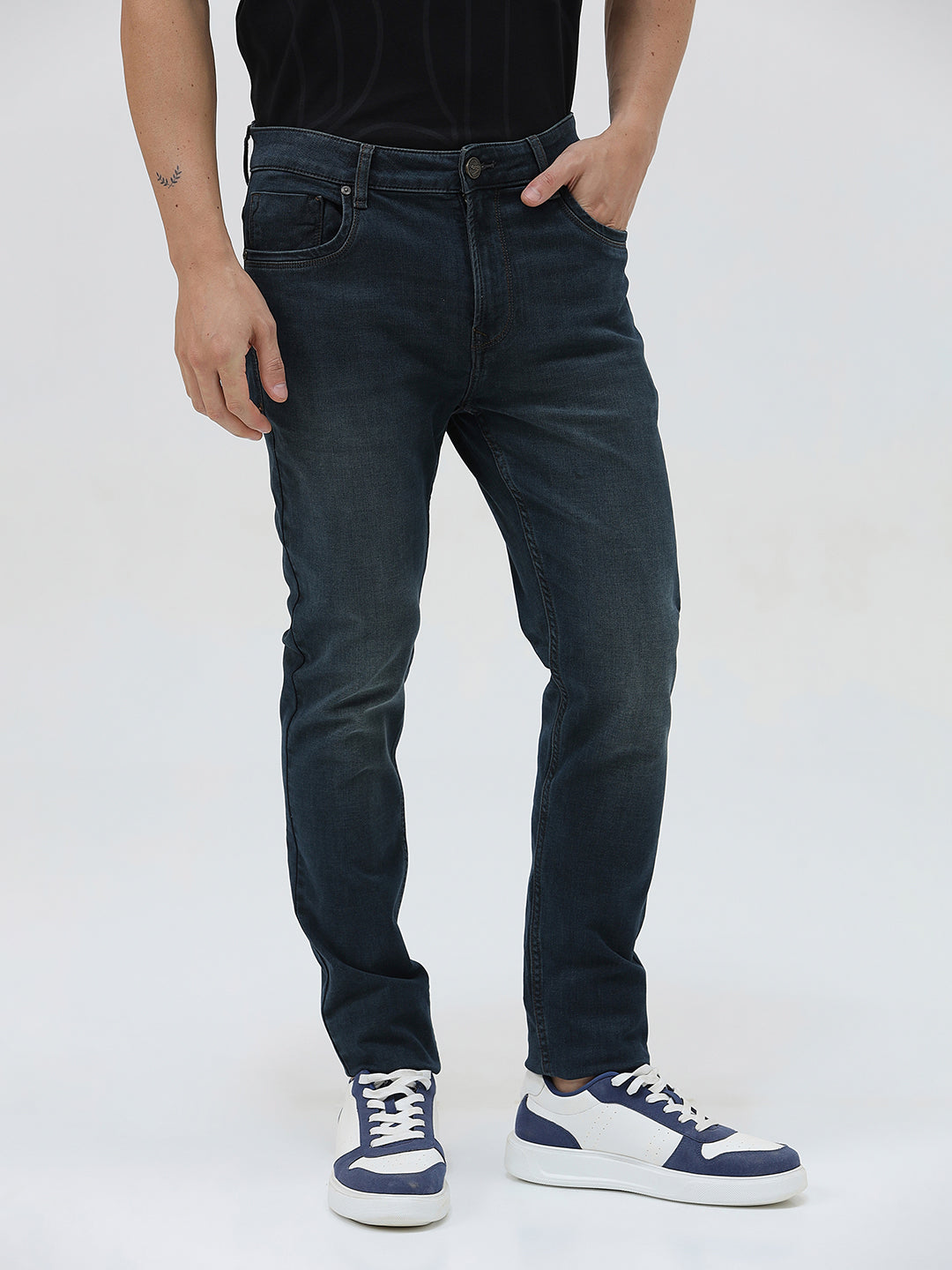 A person wearing dark blue slim-fit denim jeans, paired with a black t-shirt and navy and white sneakers, standing against a plain white studio background.