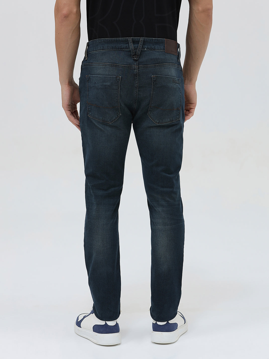 A person wearing dark blue slim-fit denim jeans, paired with a black t-shirt and navy and white sneakers, standing against a plain white studio background.