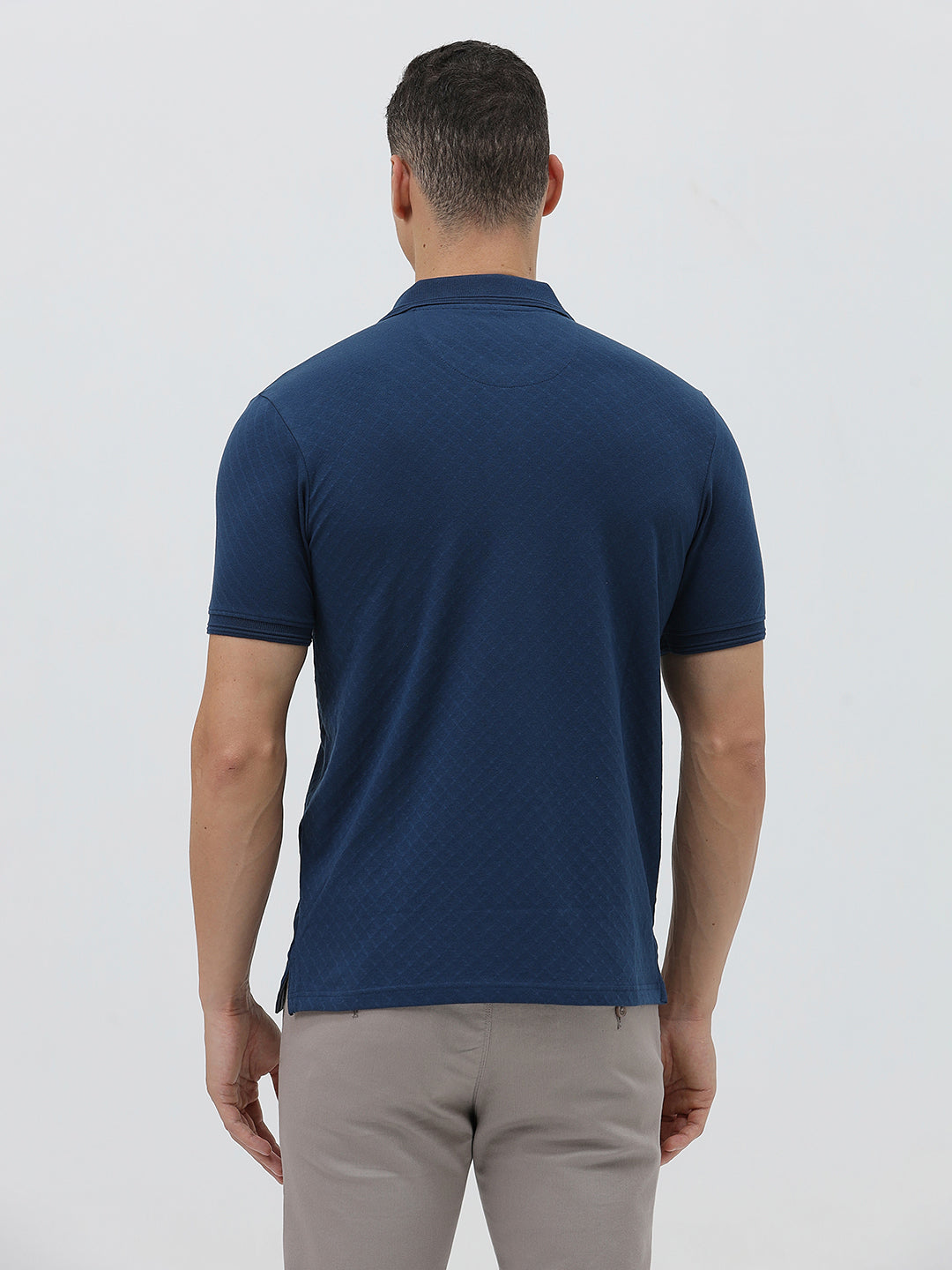 A man wearing a navy blue short-sleeve polo shirt with a chest pocket, paired with light gray trousers, standing against a plain light gray studio background.