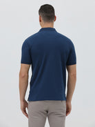 A man wearing a navy blue short-sleeve polo shirt with a chest pocket, paired with light gray trousers, standing against a plain light gray studio background.