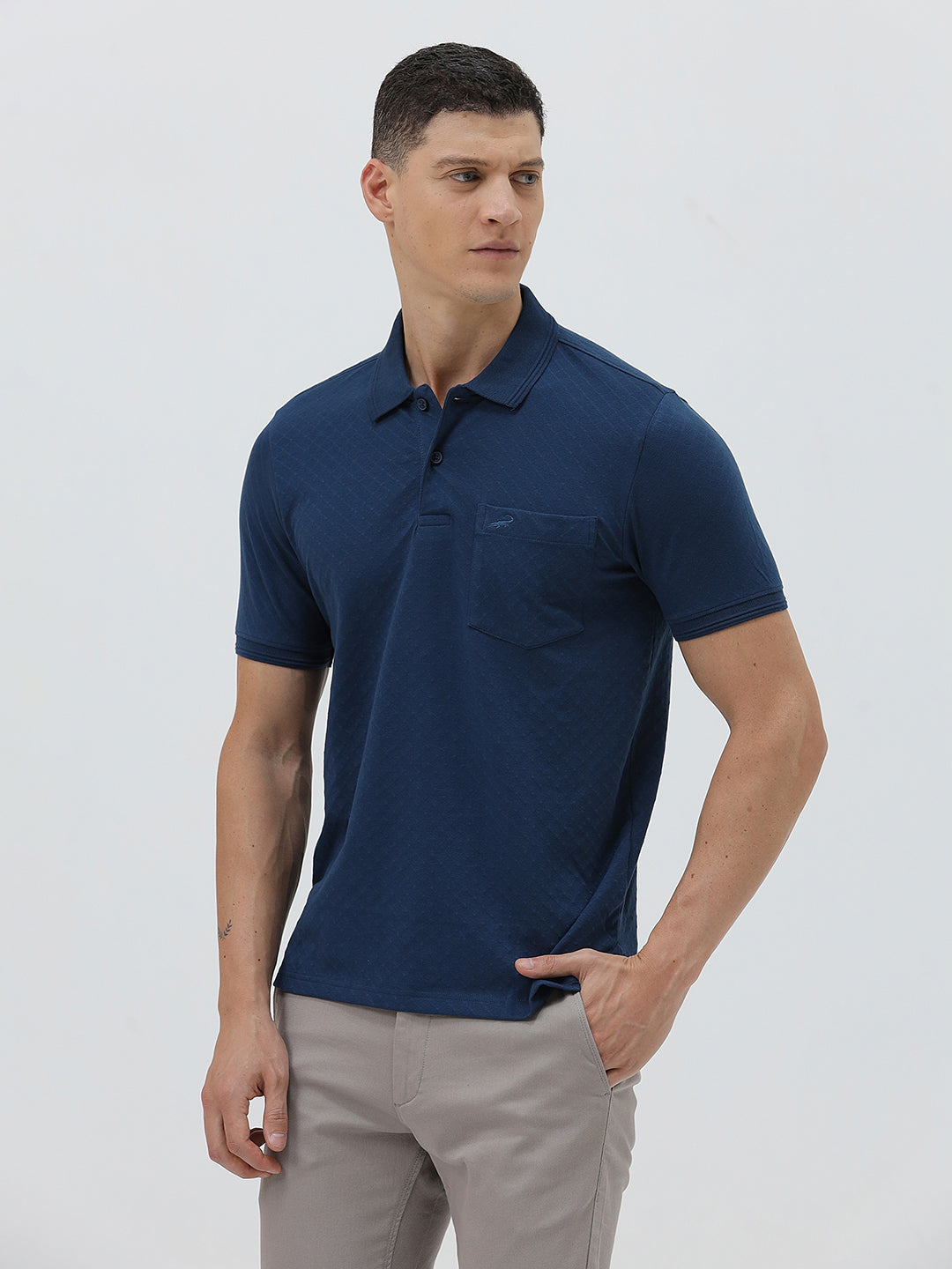 A man wearing a navy blue short-sleeve polo shirt with a chest pocket, paired with light gray trousers, standing against a plain light gray studio background.