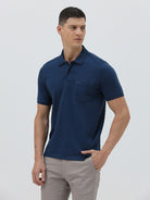 A man wearing a navy blue short-sleeve polo shirt with a chest pocket, paired with light gray trousers, standing against a plain light gray studio background.