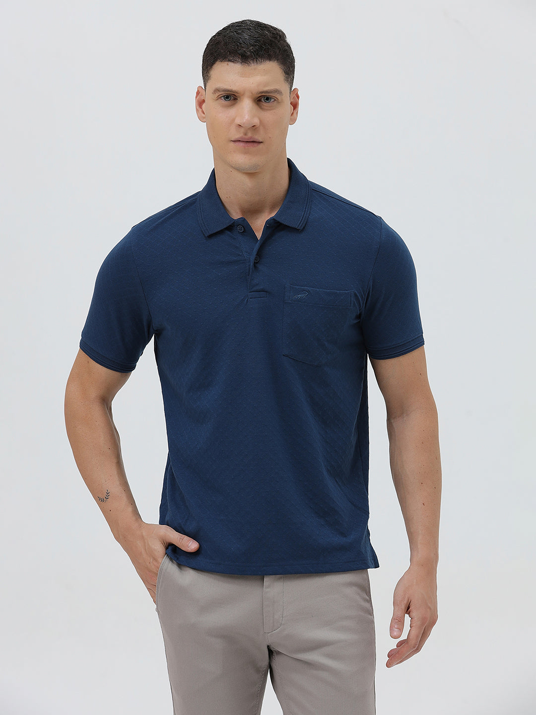 A man wearing a navy blue short-sleeve polo shirt with a chest pocket, paired with light gray trousers, standing against a plain light gray studio background.