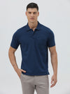 A man wearing a navy blue short-sleeve polo shirt with a chest pocket, paired with light gray trousers, standing against a plain light gray studio background.