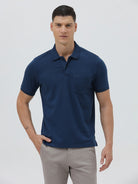 A man wearing a navy blue short-sleeve polo shirt with a chest pocket, paired with light gray trousers, standing against a plain light gray studio background.
