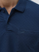 A man wearing a navy blue short-sleeve polo shirt with a chest pocket, paired with light gray trousers, standing against a plain light gray studio background.