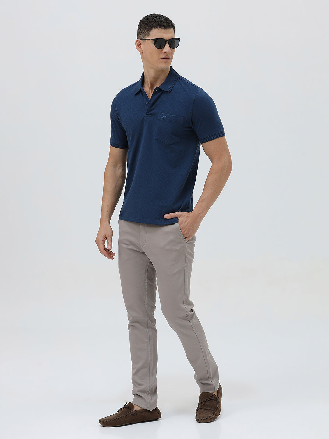 A man wearing a navy blue short-sleeve polo shirt with a chest pocket, paired with light gray trousers, standing against a plain light gray studio background.