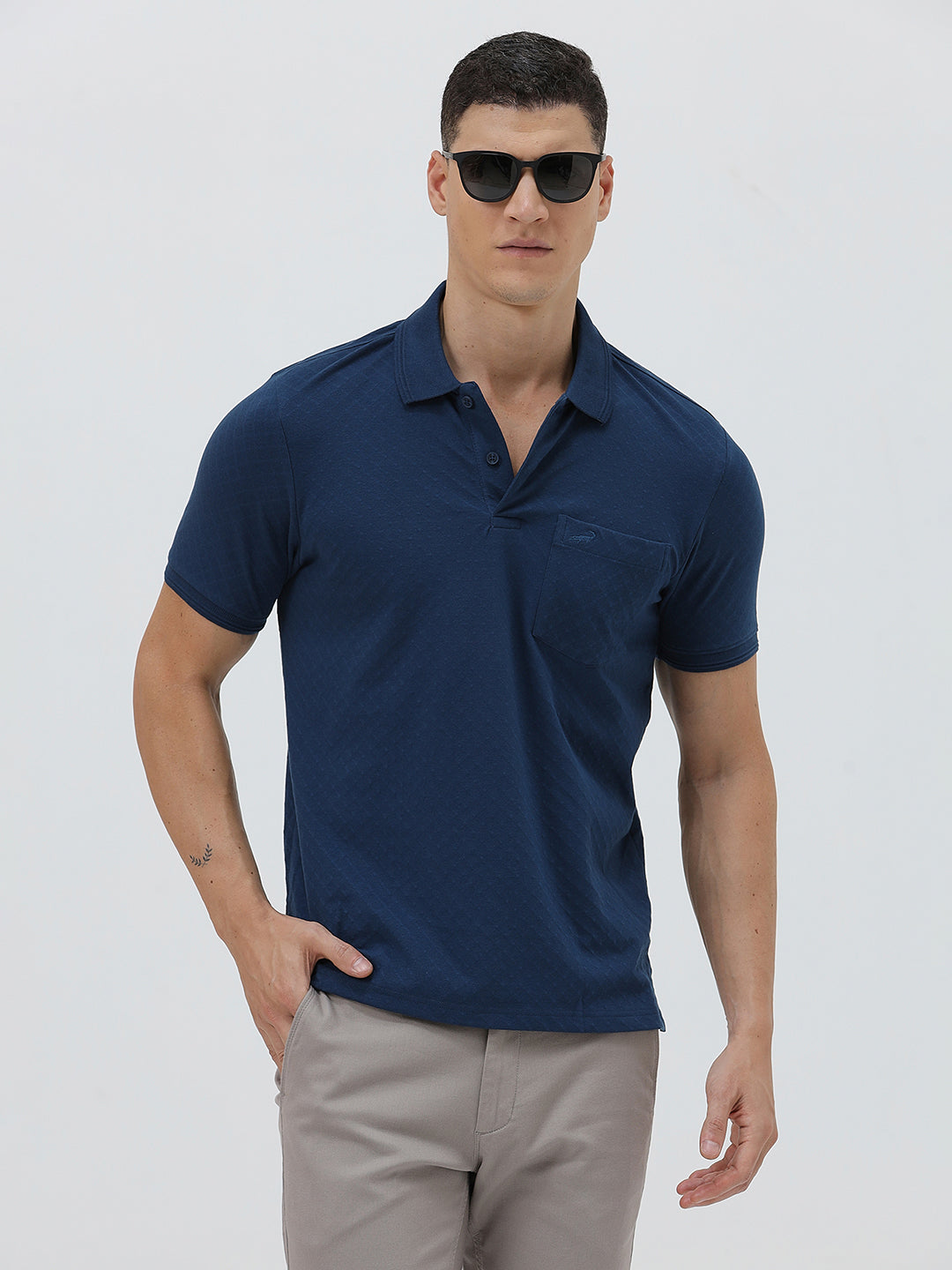 A man wearing a navy blue short-sleeve polo shirt with a chest pocket, paired with light gray trousers, standing against a plain light gray studio background.
