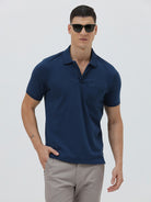 A man wearing a navy blue short-sleeve polo shirt with a chest pocket, paired with light gray trousers, standing against a plain light gray studio background.