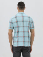 A man wearing a light blue short-sleeve polo shirt with a red and gray plaid pattern, paired with dark gray jeans, standing against a light gray studio background.