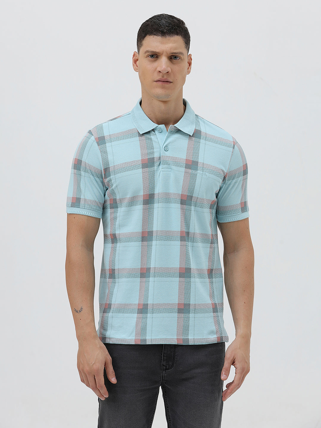 A man wearing a light blue short-sleeve polo shirt with a red and gray plaid pattern, paired with dark gray jeans, standing against a light gray studio background.