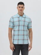 A man wearing a light blue short-sleeve polo shirt with a red and gray plaid pattern, paired with dark gray jeans, standing against a light gray studio background.