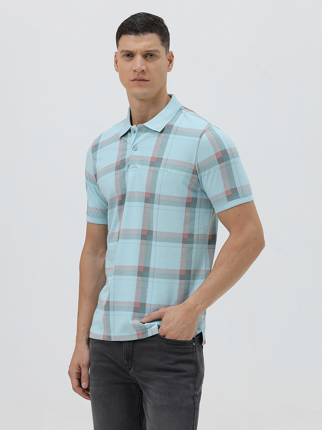 A man wearing a light blue short-sleeve polo shirt with a red and gray plaid pattern, paired with dark gray jeans, standing against a light gray studio background.