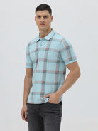 A man wearing a light blue short-sleeve polo shirt with a red and gray plaid pattern, paired with dark gray jeans, standing against a light gray studio background.