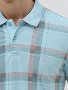 A man wearing a light blue short-sleeve polo shirt with a red and gray plaid pattern, paired with dark gray jeans, standing against a light gray studio background.