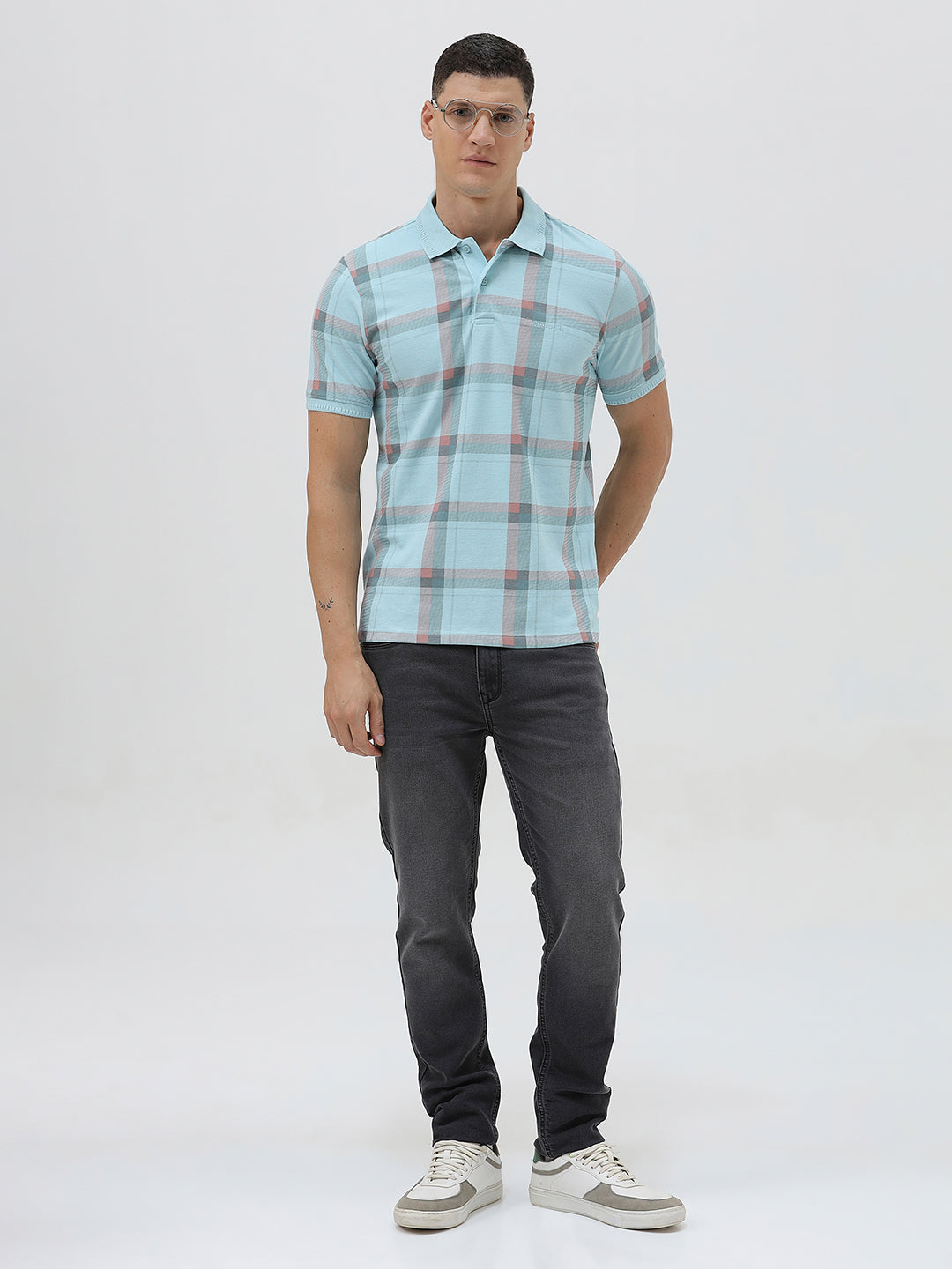 A man wearing a light blue short-sleeve polo shirt with a red and gray plaid pattern, paired with dark gray jeans, standing against a light gray studio background.